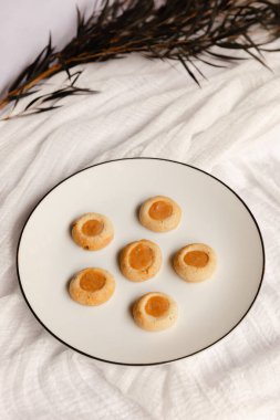 Altın kayısı merkezli altı küçük kurabiye güzel bir şekilde beyaz bir tabağa konur. Kırsal alan yumuşak kumaşlar ve biraz yeşillik içeriyor, rahatlatıcı bir atmosfer yaratıyor..