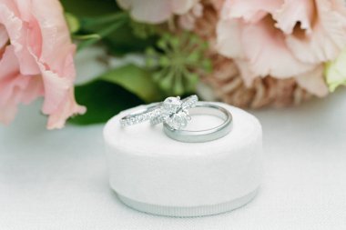 Karmaşık elmas nişan yüzüğü ve basit alyans zarif bir şekilde pembe çiçeklerle çevrili bir mindere oturur..