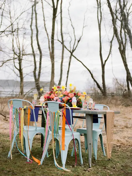 Canlı çiçekler ve renkli kurdelelerle süslenmiş güzel bir açık hava masası çıplak ağaçlar ve bulutlu gökyüzü ile çevrili huzurlu bir yerde oturur..