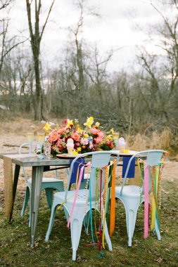 Büyüleyici bir açık hava masası canlı çiçek düzenlemeleri ve renkli dekorasyonlarla süslenir. Neşeli bir atmosferi ön plana çıkarır. Kurulumda sandalyeler ve sakin bir doğal fon yer alıyor..