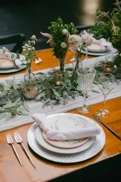 Güzelce düzenlenmiş bir yemek masası yumuşak pembe tabaklar, kristal cam eşyalar ve çiçek süslemeleri ile yemyeşil bir ortam oluşturur..