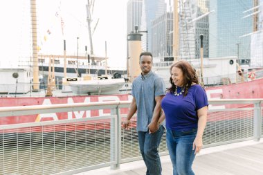 Bir çift, arka planda eski bir gemi ve modern bir şehrin siluetinin yakınında el ele tutuşurken rahat bir gezintinin tadını çıkarıyor..