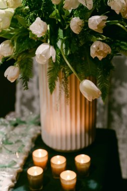 Parlayan mumlarla çevrili, dokuma vazodaki beyaz lalelerin şık düzenlemesi, sıcak bir atmosfer yaratıyor..