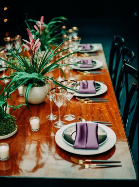 Güzel düzenlenmiş bir yemek masasında bir etkinlik için mor peçeteler, zarif tabaklar ve çiçekli süsler bulunur..