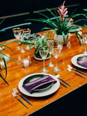 Güzel bir şekilde düzenlenmiş yemek masasında mor peçeteler, cam eşyalar ve davetkar bir atmosfer için dekoratif bitkiler bulunur..
