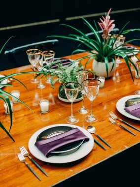 Güzel düzenlenmiş yemek masasında mor peçeteler, cam eşyalar ve canlı bitkiler var. Akşam etkinliği için mükemmel..
