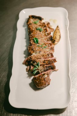 Lezzetli ızgara et, beyaz bir tepside taze otlu garnitür ve yanında bir dilim limonla..