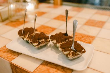 Üç porsiyon tiramisu ve sıcak bir restoran ortamında kaşık desenli bir masada sergileniyor..