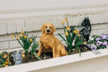 Golden Retriever heykeli, büyüleyici bir bahçe ortamında canlı çiçekler ve dekoratif elementlerin arasında bulunur..