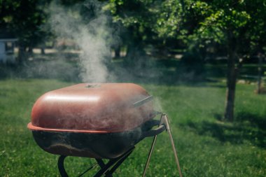 Bir barbekü ızgarası yemyeşil bir arka bahçede duman yayar. Bu da hoş bir günde yemek pişirme aktivitesi olduğunu gösterir..