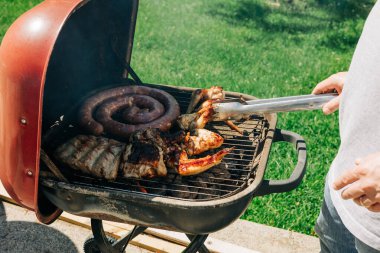 Bir insan güneşin altında çeşitli etlerle dolu barbekü ızgarasına eğilimli, canlı bir açık hava yaratıyor..