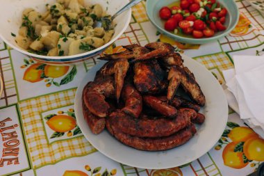 Taze domates salatası ve makarna salatasıyla birlikte çeşitli ızgara sosis ve tavuk kanatları..