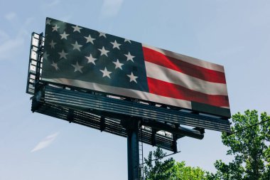 Dijital bir reklam panosunda, bir kentsel alanda açık mavi gökyüzü üzerinde belirgin bir Amerikan bayrağı yer alıyor..