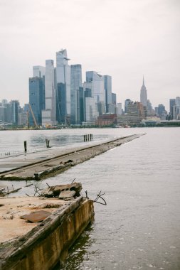 Bulutlu bir günde modern binalarla New York şehrinin ufuk çizgisi ve eski ve bakımsız bir rıhtımın yakınında ikonik bir kule görünüyor..