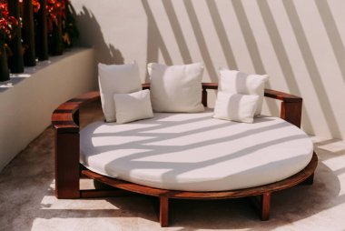 Yumuşak yastıklarla süslenmiş yuvarlak bir şezlong, dekoratif bitkiler ve gölgelerle çevrili güneş ışığıyla yıkanır..