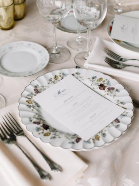 Güzel düzenlenmiş bir yemek masasında çiçek porselenleri, cilalanmış çatal bıçaklar ve misafirler için zarif bir şekilde sergilenen bir menü bulunur..