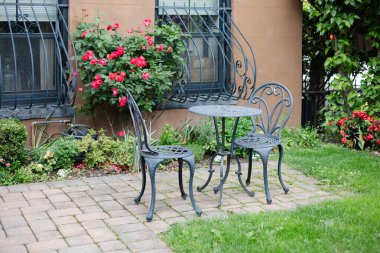 Konforlu bahçe alanında demir bir masa ve renkli çiçekler ve yeşilliklerle çevrili sandalyeler yer alıyor..