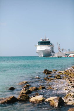 Büyük bir yolcu gemisi, güneşli bir günde, kayalık bir kıyı şeridi ve berrak sularıyla kıyıya yanaşır..