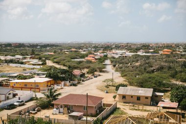 Parlak ve sıcak bir günde, ön planda çeşitli evler ve yemyeşil bitkilerle dolu canlı bir yerleşim alanı sergileniyor..