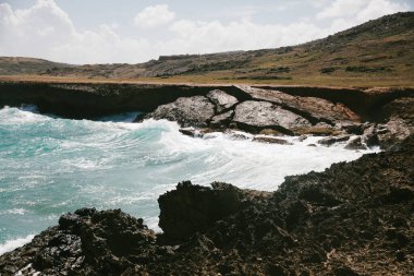 Güçlü dalgalar kayalık kıyı şeridini vurur, yeşil tepeler ve mavi gökyüzü üzerinde köpüklü beyaz dalgalar yaratır..