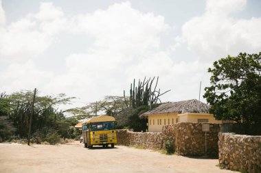Sarı bir otobüs taştan duvarlar ve bitki örtüsünden geçer kırsal bir ortamda, güneşli bir günde bulutlarla çevrili..