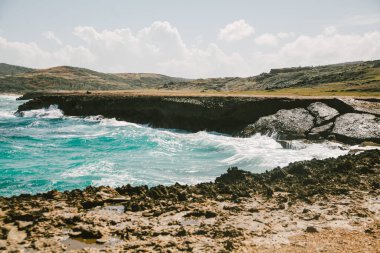 Sakin dalgalar dalgalanarak dalgalanır kayalık kıyılarda Tepeler arka planda berrak mavi gökyüzünün altında.