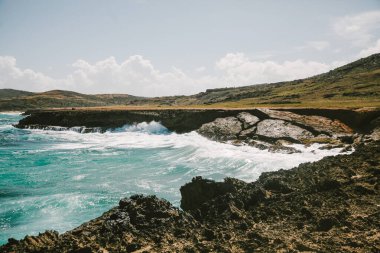 Turkuaz dalgalar engebeli kayalara karşı kırılıyor, arka planda gür tepeler berrak bir gökyüzünün altında.