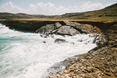 Güçlü dalgalar arka planda dağınık bulutlar ile berrak mavi bir gökyüzü altında kıyı boyunca kayalarla etkileşime girer..