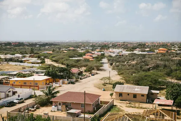Parlak ve sıcak bir günde, ön planda çeşitli evler ve yemyeşil bitkilerle dolu canlı bir yerleşim alanı sergileniyor..