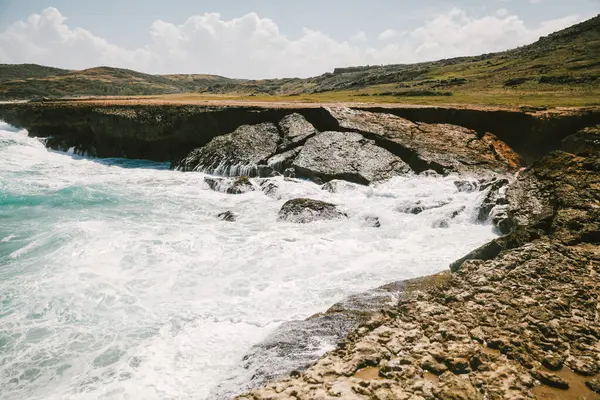 Güçlü dalgalar arka planda dağınık bulutlar ile berrak mavi bir gökyüzü altında kıyı boyunca kayalarla etkileşime girer..