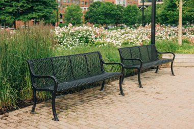 İki siyah metal sırık, yeşil yapraklar ve açık mavi gökyüzünün altında renkli çiçeklerle çerçevelenmiş tuğla bir yolda oturur..