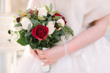 Bir gelin, serin bir kış günü, yemyeşil aksanlı kırmızı ve beyaz bir gül buketini zarifçe tutar..