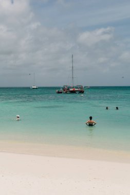 Sıcak plaj günü, bulutlu bir gökyüzünün altında bir yelkenli dinlenirken insanların berrak sularda yüzdüğü..