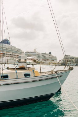 Küçük bir yelkenli, bulutlu bir günde limandaki büyük yolcu gemilerinin yanında demirler..