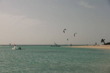 Uçurtma sörfçüleri turkuaz sularda süzülürken sahile gidenler bulutlu bir gökyüzünün altında dinleniyorlar..