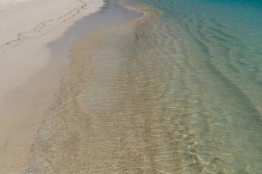 Saf bir kıyı şeridinde hafif dalgalar, berrak mavi suları ve ılık bir günde yumuşak bej kumu sergiliyor..