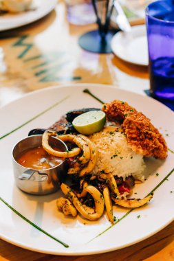 Renkli tabakta ızgara kalamar, kızarmış balık, pirinç ve canlı bir yemek ortamında limon dilimi yer alıyor..