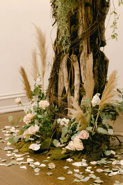 Kapalı mekanda loş ışıklandırılmış bir ağacın dibinde gül ve yeşillik içeren zarif bir çiçek aranjmanı.