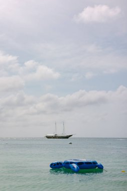 Mavi bir sal bulutlu bir gökyüzünün altında durgun suda yüzerken, bir yelkenli gemi uzak bir yere demir atar..