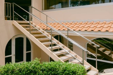 Paslanmaz çelik parmaklıklı merdiven, yeşil çalılıklarla çevrili modern bir binanın üst katına çıkıyor..