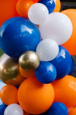 Mavi, turuncu, beyaz ve altın renkli canlı balon kümeleri açık havada bir kutlama ortamı oluşturur.