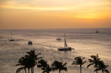 Güneş ufukta batarken, sakin sularda süzülen yelkenliler sakin bir sahil atmosferi yaratıyor..