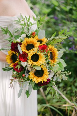 Bir gelin, bahçesinde yeşilliklerle çevrili ayçiçekleri ve kırmızı güllerle dolu canlı bir buket tutar..