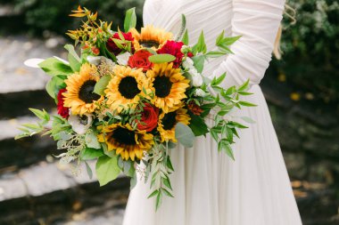 Bir gelin, açık havada süzülen beyaz bir elbise giyerken ayçiçeği ve güllerle dolu canlı bir buket tutar..