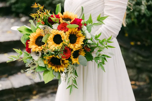 Bir gelin, açık havada süzülen beyaz bir elbise giyerken ayçiçeği ve güllerle dolu canlı bir buket tutar..
