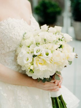 Bir gelin, gündüz vakti, sakin bir bahçede dikilirken bir buket beyaz çiçek tutar..