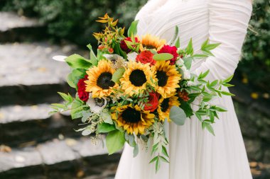 Bir gelin rengarenk bir demet ayçiçeği tutar ve bir bahçede dikilirken çeşitli çiçekler açar..