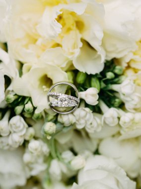 Alyanslar zarif bir şekilde yumuşak beyaz çiçeklerin arasına yerleştirilmiştir. Aşkı ve birliği sembolize eder..