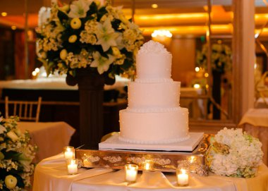 Göz kamaştırıcı üç katlı düğün pastası çiçeklerle ve mum ışığıyla çevrili süslü bir masada oturur..