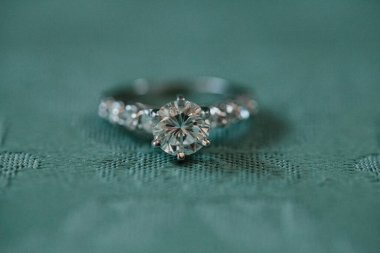 Güzel elmas yüzük yuvarlak kesimli bir taş ve ince detaylar içeriyor, desenli bir deniz mavisi kumaşı üzerinde incelikle dinleniyor..